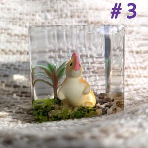 May include: A clear cube filled with a small, yellow and white figurine of a rabbit with a pink nose, green plastic plants, and small rocks. The cube is sitting on a white, textured surface.