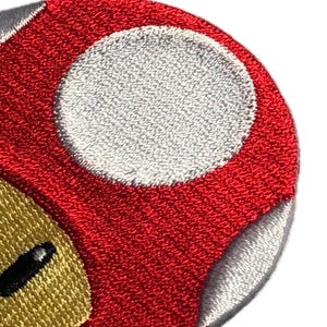 Könnte beinhalten: Gestickte Aufnäher mit einem roten Pilz mit weißen Punkten und einem grünen Pilz mit weißen Punkten. Beide haben eine beige Basis mit schwarzen ovalen Augen. Diese Aufnäher sind wahrscheinlich von einem beliebten Videospiel inspiriert.