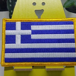 Puede incluir: Un parche bordado de la bandera griega azul y blanca con un borde dorado. El parche se muestra en un soporte amarillo.