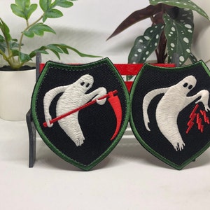 May include: Two embroidered iron-on patches featuring a white ghost figure. One patch shows the ghost holding a red scythe, the other shows the ghost holding red lightning bolts. Both patches have a black background with a green border.