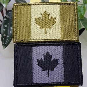 Könnte beinhalten: Zwei gestickte kanadische Flaggen-Patches. Der obere Patch ist olivgrün, beige und weiß. Der untere Patch ist schwarz, grau und weiß.