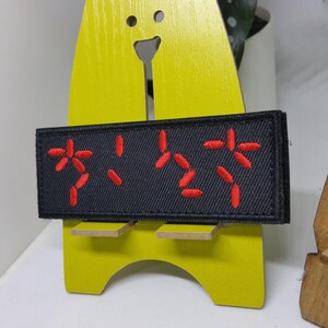Puede incluir: Un parche rectangular negro con caracteres bordados en rojo. El parche se muestra en un soporte de madera amarillo. El texto bordado parece estar en un idioma extranjero. El parche tiene un borde negro.