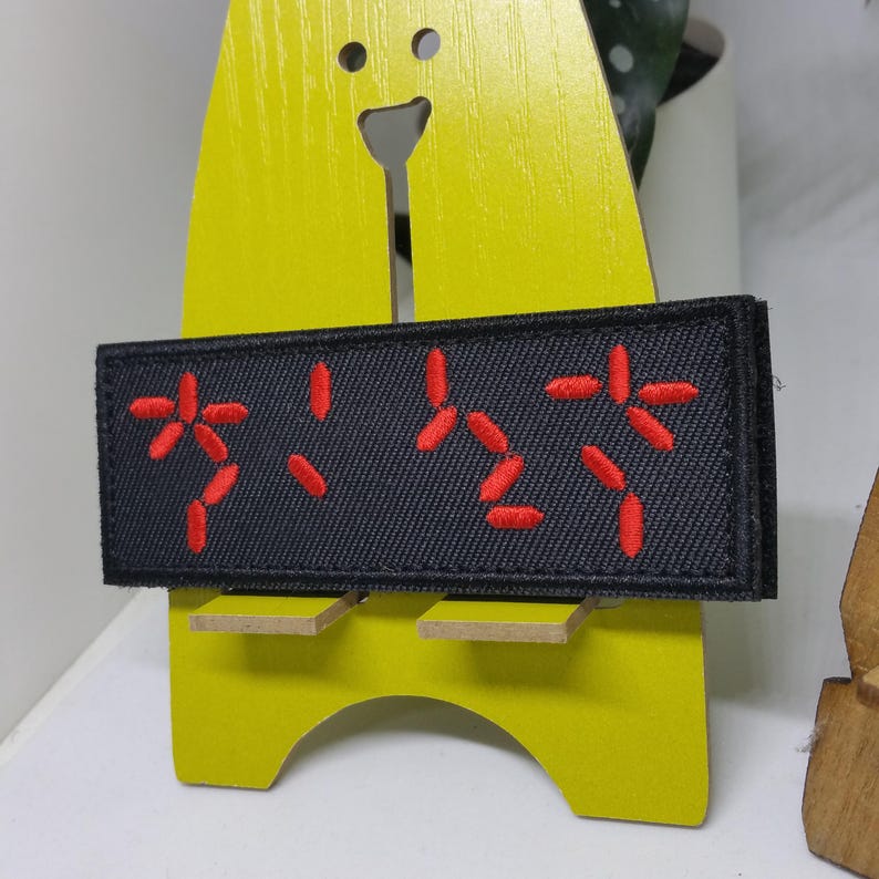 Puede incluir: Un parche rectangular negro con caracteres bordados en rojo. El parche se muestra en un soporte de madera amarillo. El texto bordado parece estar en un idioma extranjero. El parche tiene un borde negro.