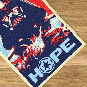 May include: Embroidered patch featuring a stylized image of Darth Vader in red and white against a navy blue background. The word "HOPE" is displayed in light blue, along with a symbol. The patch has a cream-colored border.