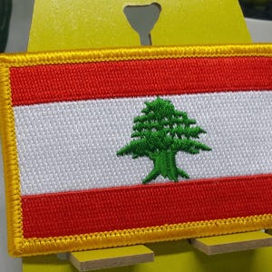 Może przedstawiać: Naszywka haftowana z flagą Libanu. Flaga jest czerwona, biała i zielona, z zielonym cedrem na białym tle.