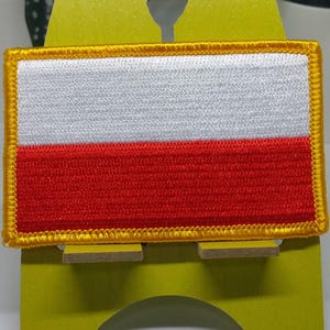 May include: A white and red embroidered patch with a gold border, depicting the flag of Poland. The patch is displayed on a yellow stand.