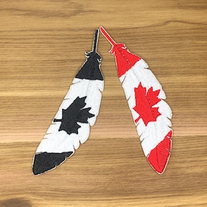 May include: Two embroidered feather patches. One is black and white with a black maple leaf, the other is red, white, and features a red maple leaf. The patches are on a wooden surface.