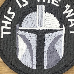 Op de afbeelding: Een cirkelvormige zwarte patch met een Mandalorian helm ontwerp in grijstinten en wit. De tekst "THIS IS THE WAY" is in het wit rond de helm geborduurd. De patch is waarschijnlijk voor kleding of accessoires.