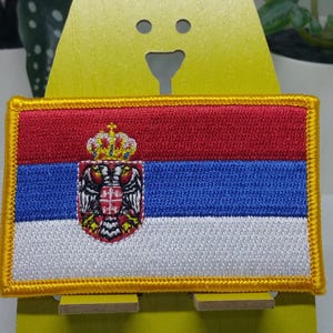 Puede incluir: Un parche bordado rojo, azul y blanco con el escudo de armas de Serbia. El parche tiene un borde dorado y se muestra en un soporte amarillo.