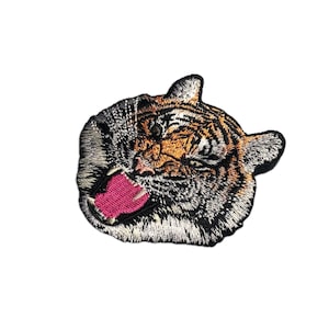 Könnte beinhalten: Gestickter Tiger-Aufnäher mit detailliertem Design eines brüllenden Tigerkopfes. Der Aufnäher zeigt orange, schwarze und silberne Fäden mit einer leuchtend rosa Zunge. Geeignet für Jacken, Taschen oder andere Stoffartikel.