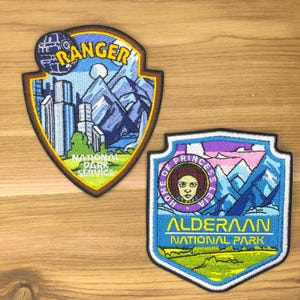 Pode incluir: Dois patches bordados com um design de escudo. Um patch apresenta uma Estrela da Morte e a palavra "Ranger" acima de uma cena de montanha e cidade. O outro patch apresenta a Princesa Leia e as palavras "Alderaan National Park".