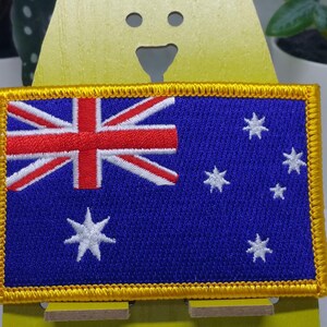 Op de afbeelding: Een geborduurde patch met de Australische vlag, in blauw en wit met een gouden rand. De patch wordt getoond op een gele standaard.