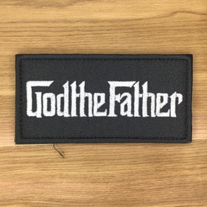 May include: A rectangular black patch with white embroidered text that reads "GodtheFather". The patch is likely for clothing or accessories, and is set against a wooden background.