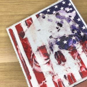 Può includere: Una toppa quadrata con un design invecchiato della bandiera americana e un teschio sovrapposto. Le strisce rosse e le stelle bianche della bandiera sono visibili, con un disegno del teschio in bianco e viola. La toppa ha un bordo bianco.
