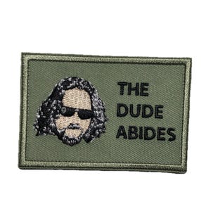 Pode incluir: Um remendo verde bordado com uma imagem de desenho animado do The Dude do filme The Big Lebowski usando óculos de sol e barba. O texto "THE DUDE ABIDES" é bordado em preto abaixo da imagem.