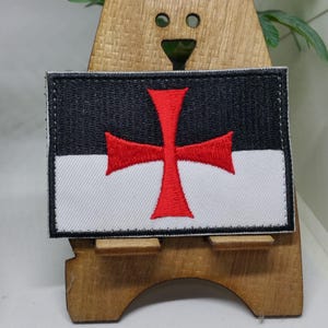 Puede incluir: Un parche bordado blanco y negro con una cruz roja. El parche est&aacute; en un soporte de madera.