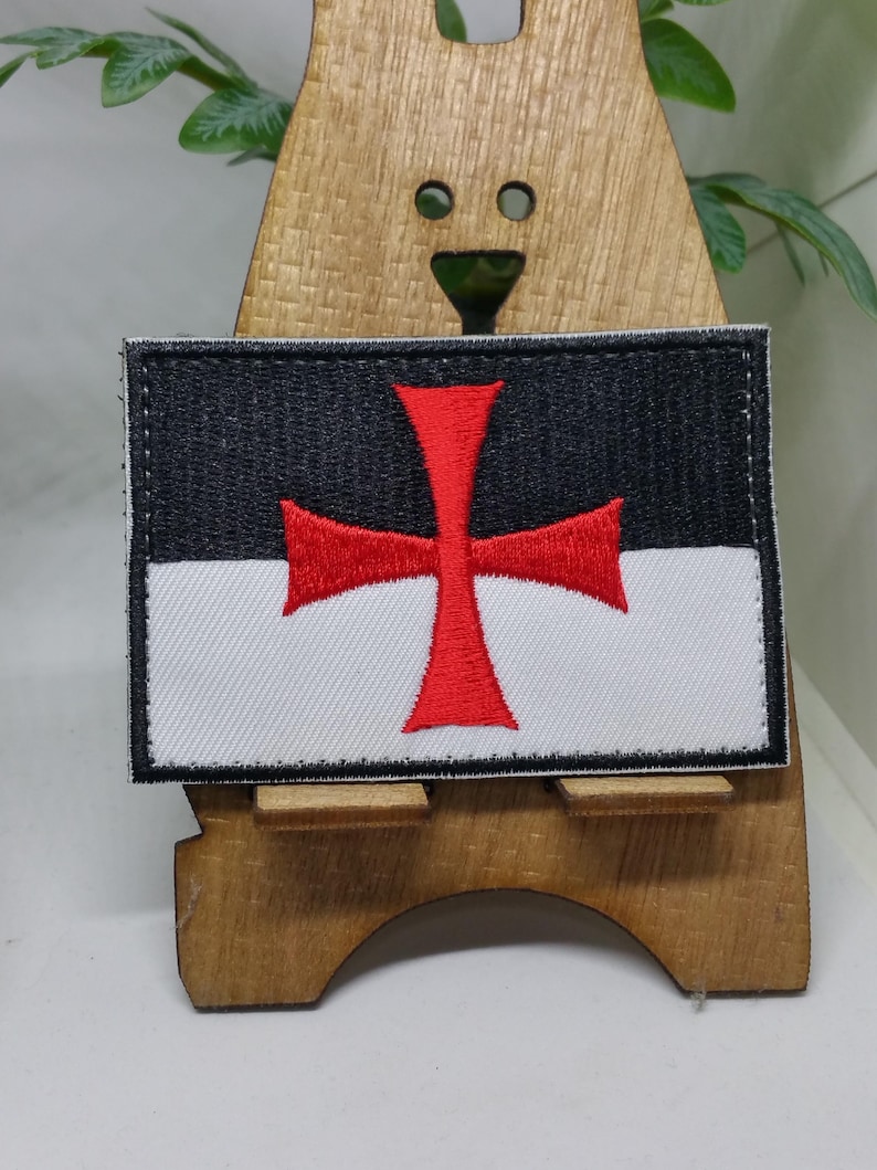 Puede incluir: Un parche bordado blanco y negro con una cruz roja. El parche est&aacute; en un soporte de madera.