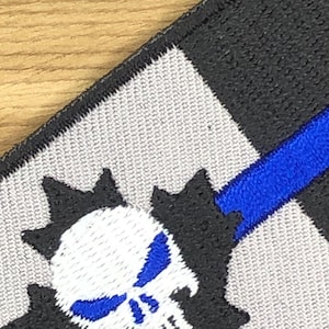 May include: Embroidered patch with a white skull and blue detailing, set on a grey and black background. A blue stripe cuts across the design. The patch has a black border and a textured finish.