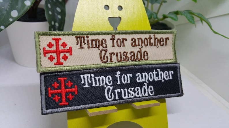Pu&ograve; includere: Due toppe ricamate con il testo "Time for another Crusade" e una croce rossa con quattro croci pi&ugrave; piccole. La toppa superiore &egrave; beige con testo marrone e un bordo verde. La toppa inferiore &egrave; nera con testo bianco e un bordo grigio.