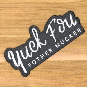Può includere: Una toppa rettangolare nera ricamata con le parole "Yuck Fou" in un carattere bianco corsivo. Sotto, il testo "FOTHER MUCKER" è in un carattere bianco più piccolo, senza grazie. La toppa ha un bordo bianco.