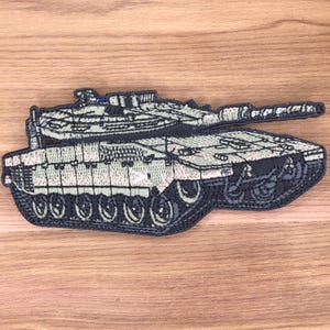 Könnte beinhalten: Gestickter Aufnäher mit einer detaillierten Darstellung eines Militärpanzers. Der Aufnäher hat ein Tarnmuster in Grün- und Beigetönen mit schwarzen Konturen. Die Ketten und der Turm des Panzers sind deutlich erkennbar.
