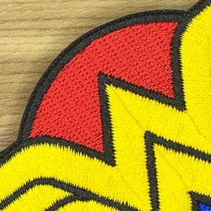 May include: Close-up of a colorful embroidered patch. The patch features a red and yellow design with black outlining. The texture of the embroidery is visible, adding depth to the design. The patch is likely for clothing or accessories.