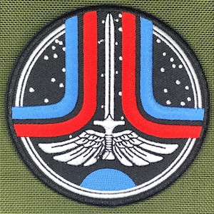 May include: Circular patch with a black backdrop, a white sword, and red and blue curved lines. A white winged emblem is at the bottom. The patch has a white border and a blue semi-circle. White dots represent stars.