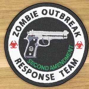 May include: Embroidered patch with a black border and a white background. The patch features a silver handgun in the center, surrounded by the words "ZOMBIE OUTBREAK RESPONSE TEAM" and "SECOND AMENDMENT" in white and green text. Red biohazard symbols are on either side.