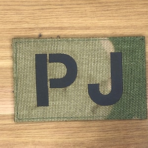 May include: A rectangular patch with the letters "PJ" in black, centered on a camouflage-patterned background. The patch has a textured, woven appearance, and the edges are neatly stitched. The background features shades of green and brown.