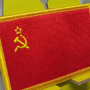 Może przedstawiać: Naszywka haftowana na czerwono z żółtą obwódką. Naszywka przedstawia żółty młot i sierp z żółtą gwiazdą nad nimi.