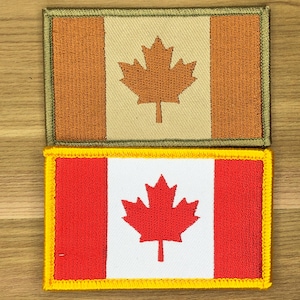 Op de afbeelding: Twee geborduurde patches met de Canadese vlag. De bovenste patch heeft een beige achtergrond met een bruin esdoornblad en een bruine rand. De onderste patch heeft een witte achtergrond met een rood esdoornblad en een rood-gele rand.