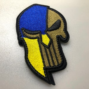 May include: An embroidered patch featuring a Spartan helmet with a skull face. The helmet is blue and yellow, outlined in black, and the skull has black eyes.