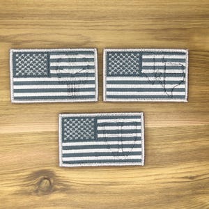 Puede incluir: Tres parches rectangulares con el diseño de la bandera estadounidense en tonos grises y blancos. Cada parche tiene una superposición gráfica diferente: una calavera, un contorno estatal y un escudo. Los parches se muestran sobre una superficie de madera.