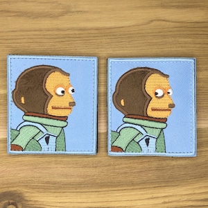 May include: Two embroidered patches depicting a monkey's side profile with a surprised expression. The monkey has brown fur, a tan face, and wears a green jacket. The patches are set against a light blue backdrop.