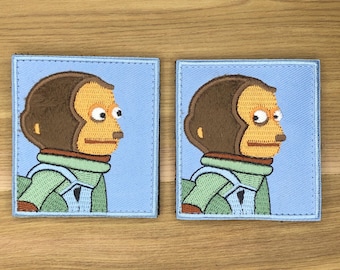 May include: Two embroidered patches depicting a monkey's side profile with a surprised expression. The monkey has brown fur, a tan face, and wears a green jacket. The patches are set against a light blue backdrop.