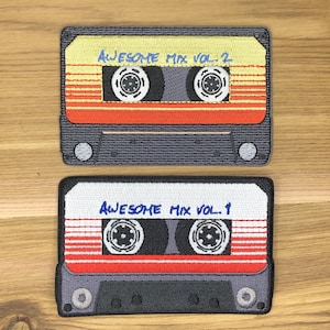 May include: Two embroidered patches shaped like cassette tapes. The top patch is yellow, red, and gray, with the text "AWESOME MIX VOL. 2." The bottom patch is white, red, and gray, with the text "AWESOME MIX VOL. 1."