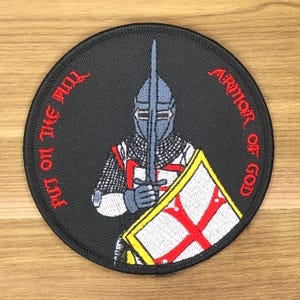 May include: Round black patch featuring a knight in armor holding a sword and shield. The shield has a red cross on a white background. Text around the edge reads "PUT ON THE FULL ARMOR OF GOD" in red.