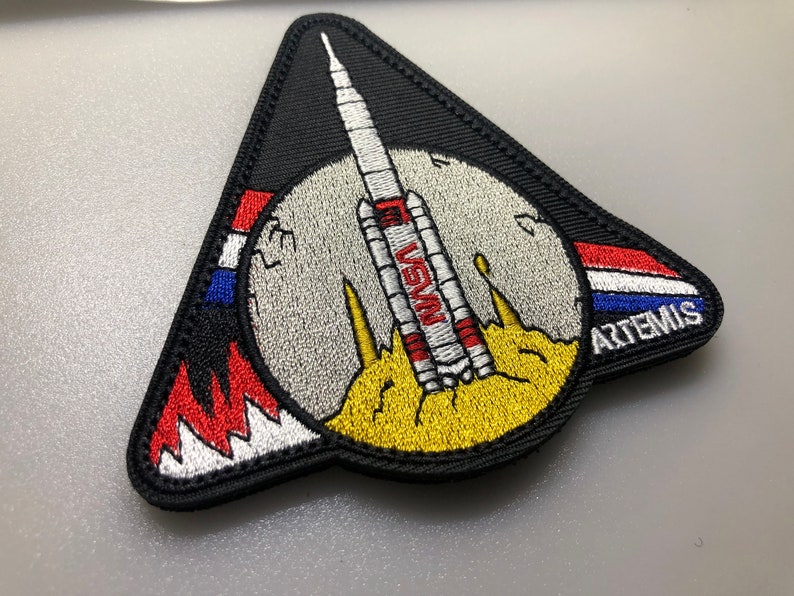 Puede incluir: Un parche triangular negro con un cohete blanco y rojo lanz&aacute;ndose hacia una luna gris con el texto "NASA" y "ARTEMIS" bordado en &eacute;l.
