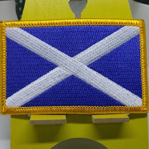May include: A blue and white Scottish flag patch with a gold border. The patch is embroidered and has a stitched edge.