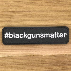 Può includere: Una toppa rettangolare nera con il testo bianco "#blackgunsmatter". La toppa ha angoli arrotondati ed è realizzata in un materiale strutturato. Il testo è in un font sans-serif.