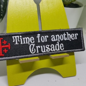 Pu&ograve; includere: Toppa ricamata nera con una croce rossa e il testo "Time for another Crusade" in bianco.