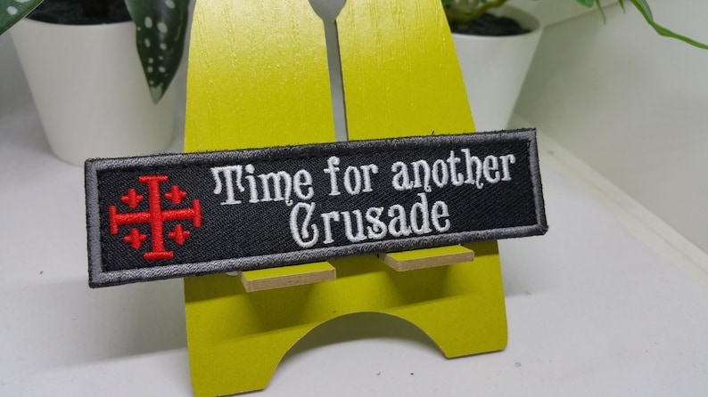Pu&ograve; includere: Toppa ricamata nera con una croce rossa e il testo "Time for another Crusade" in bianco.