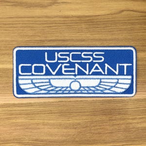 Op de afbeelding: Een rechthoekige patch met een blauwe achtergrond en witte rand. De tekst "USCSS COVENANT" wordt weergegeven in witte blokletters. Daaronder bevindt zich een witte afbeelding van gestileerde vleugels en een centrale cirkel.