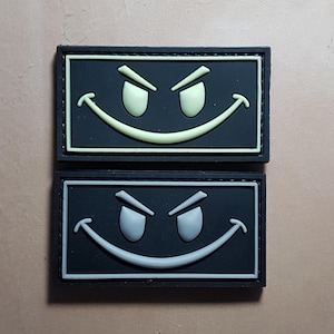 Puede incluir: Dos parches rectangulares negros con un contorno blanco. Cada parche presenta una cara sonriente blanca con las cejas levantadas. El parche superior tiene una cara sonriente verde claro, mientras que el parche inferior tiene una cara sonriente gris.