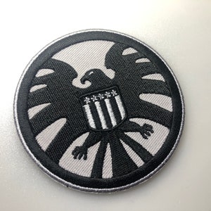 May include: A black and white embroidered patch featuring the S.H.I.E.L.D. logo. The logo is a stylized eagle with a shield on its chest. The shield has a white background with five red stars and five white stripes.