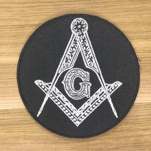 May include: A circular, black patch displaying a white Masonic emblem. The design incorporates a compass, a set square, and the letter "G" at the centre. The patch is likely intended for clothing or accessories.