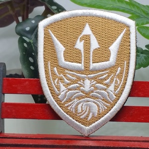 Puede incluir: Un parche bordado de color marrón claro con un tridente blanco y una cara estilizada en el centro. El parche tiene forma de escudo.