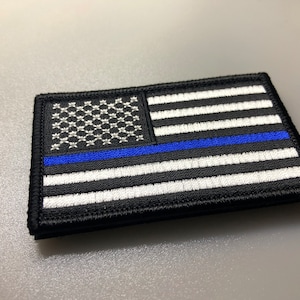 Peut inclure: Un patch de drapeau américain noir et blanc avec une fine ligne bleue au milieu. Le patch est brodé et a une bordure noire.