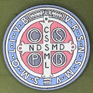 Könnte beinhalten: Runder, gestickter Aufnäher mit einem Kreuzdesign und verschiedenen Symbolen und Buchstaben. Der Aufnäher hat einen hellblauen Rand mit weißen Buchstaben, einen rosa Innenkreis und ein weißes Kreuz. Das Wort "PAX" steht oben.