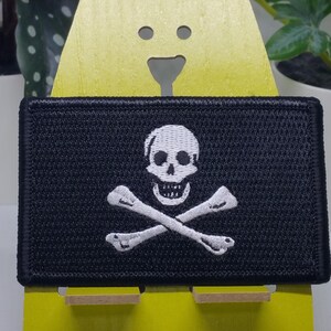 May include: A black embroidered patch with a white skull and crossbones design. The patch is displayed on a yellow stand.